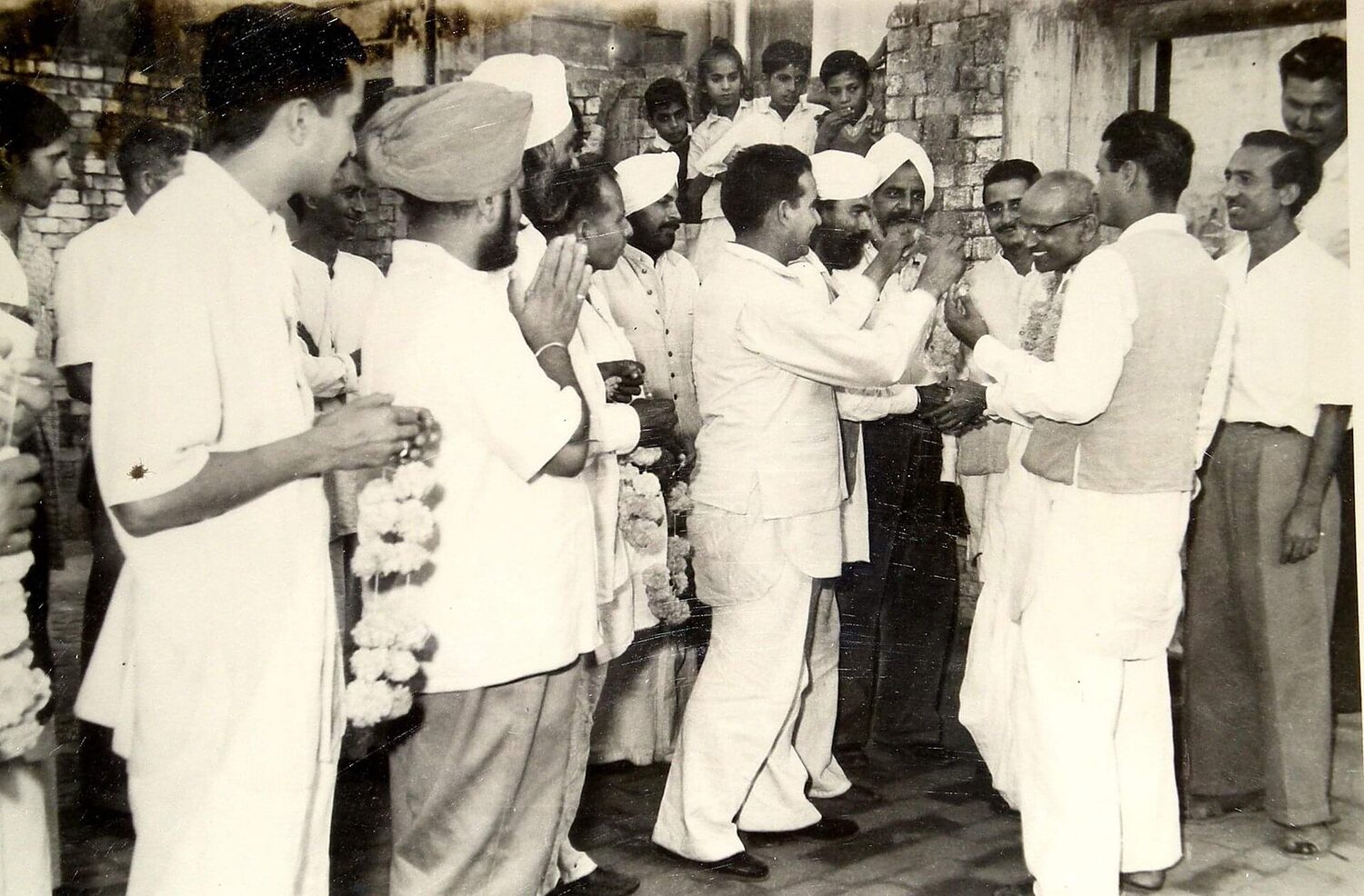 A crowd of people in white clothing gathered outdoors to garland Batukeshwar Dutt, who stands at the right with folded hands.